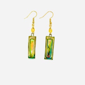 Peut inclure: Une paire de boucles d'oreilles faites à la main avec des crochets d'oreille dorés. Chaque boucle d'oreille présente un cadre rectangulaire de couleur bronze avec un motif abstrait coloré dans les tons de vert, de jaune et d'orange. De petites perles jaunes ornent le haut de chaque boucle d'oreille.
