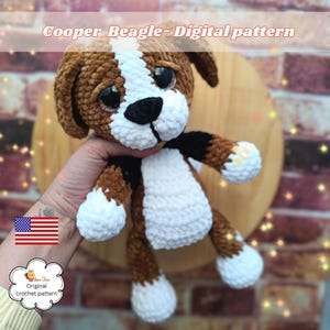 Puede incluir: Un juguete de perro beagle tejido a ganchillo con hilo marrón, blanco y negro, con grandes ojos oscuros y una nariz negra. El texto Cooper Beagle- Digital pattern es visible en la parte superior y un pequeño logo con el texto Original crochet pattern está debajo.