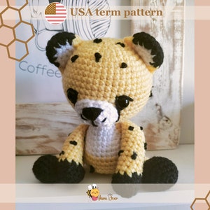 Pode incluir: Um brinquedo de amigurumi de guepardo de crochê com manchas pretas, orelhas pretas e pés pretos. O brinquedo está sentado em uma superfície de madeira.