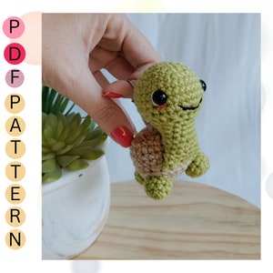May include: A green crocheted turtle with a brown shell and black eyes. The turtle is being held by a hand.
