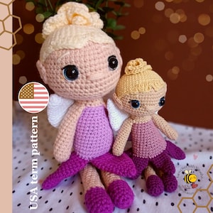 Pode incluir: Duas bonecas de fada de crochê, uma maior que a outra, ambas com pele rosa, cabelo loiro, vestidos roxos e asas brancas. As bonecas estão sentadas em um fundo branco com um padrão de pequenos pontos pretos.