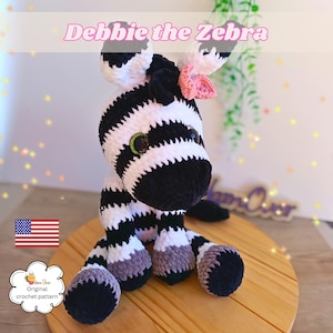 Puede incluir: Un peluche de cebra tejido a ganchillo llamado Debbie the Zebra con rayas blancas y negras, una melena negra y una pequeña flor rosa como accesorio en la cabeza. La cebra tiene grandes ojos oscuros y pezuñas grises, hecha de hilo suave.