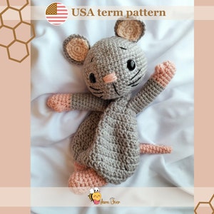 Puede incluir: Un juguete de ratón de crochet gris con detalles en rosa. El ratón tiene una cola larga y está acostado sobre una superficie blanca. El juguete está hecho con un hilo texturizado y tiene una expresión linda y amigable.
