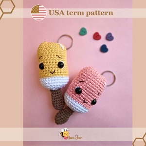 Puede incluir: Dos llaveros de paleta de helado de crochet, uno amarillo y otro rosa, con caras sonrientes y mangos de palo marrón. Los llaveros están sobre un fondo rosa con pequeños botones en forma de corazón.