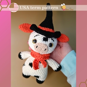 Può includere: Una mucca di Halloween all'uncinetto che indossa un cappello da strega nero con un bordo arancione e una sciarpa arancione. La mucca ha macchie nere e grandi occhi marroni.