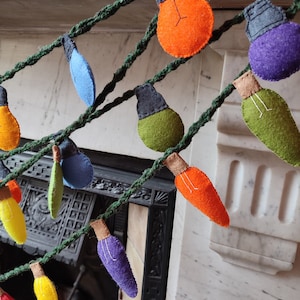 May include: A vibrant felt light bulb garland strung on a green rope. The garland showcases an array of coloured bulbs, such as orange, purple, green, blue, and yellow. Each bulb has a grey cap and a brown base. A decorative item.