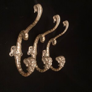 Puede incluir: Tres perchas de pared de metal plateado con un diseño único. Cada gancho presenta una parte superior en forma de cobra y una cara decorativa en la base. Los ganchos están montados sobre un fondo negro.