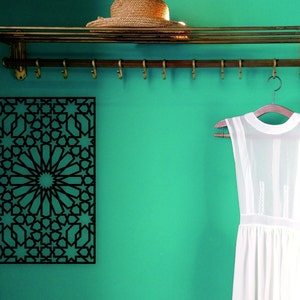 May include: A black metal wall decor with a geometric pattern of stars and circles. The decor is hanging on a teal wall with a brass shelf and hooks above it. A white dress is hanging on a wooden hanger on the hooks.