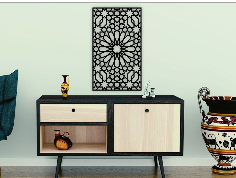 May include: A wooden cabinet with two drawers and a shelf. The cabinet is light wood with black trim. There is a black metal wall decor with a geometric pattern above the cabinet. There are two small vases on the top of the cabinet, one is brown and the other is gold. There is a large vase with a black and white pattern on the right side of the cabinet.