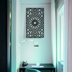 May include: A black metal wall decor with a geometric pattern of stars and circles. The decor is hanging on a white wall in a room with a desk and a window.