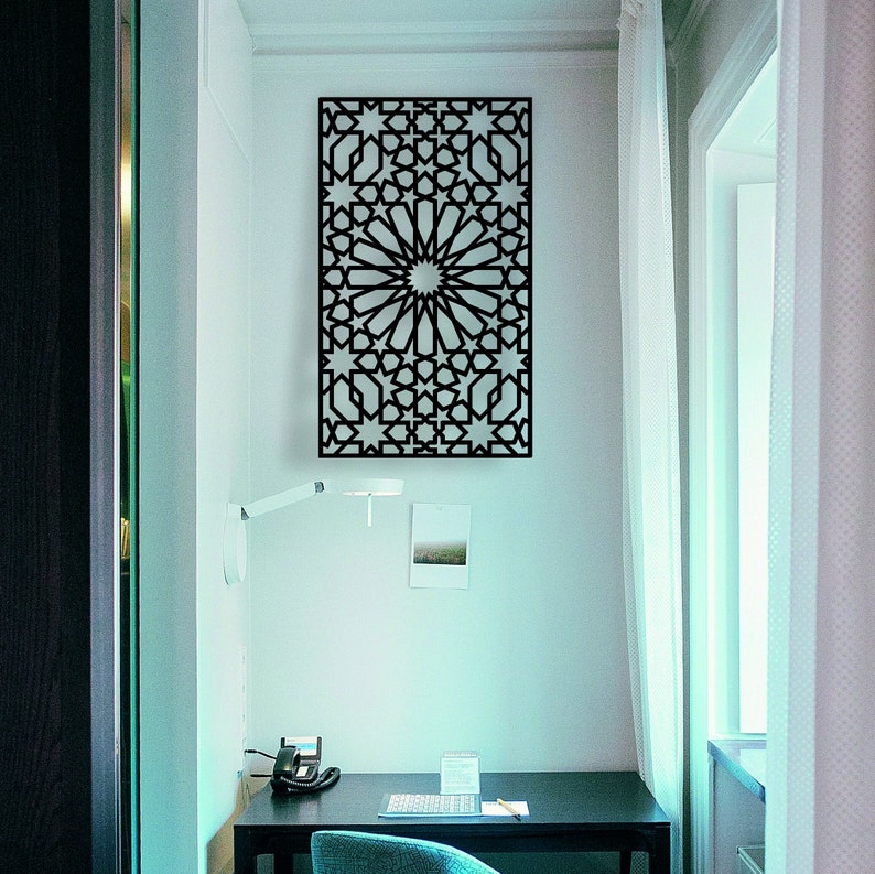 May include: A black metal wall decor with a geometric pattern of stars and circles. The decor is hanging on a white wall in a room with a desk and a window.