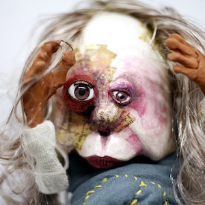 Peut inclure: Une poupée faite à la main avec un visage unique et expressif. La poupée présente un mélange de textures et de couleurs, avec un accent sur les yeux et la bouche. Les cheveux de la poupée sont longs et gris, et elle porte un vêtement bleu avec des coutures jaunes.