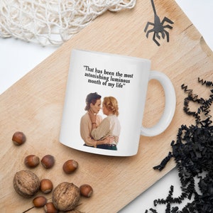 May include: White ceramic mug featuring a vintage image of two women embracing, with the quote "That has been the most astonishing luminous month of my life." The mug has a curved handle and is set on a wooden surface with nuts.