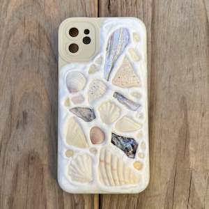 May include: A white iPhone case with a mosaic design made of seashells. The case has a cutout for the camera lens and the text "Pro Camera" is printed on the case.
