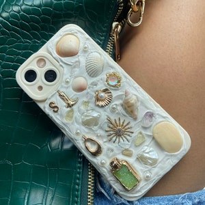 May include: White iPhone case with a seashell and pearl design. The case has a variety of seashells, pearls, and other beach-themed embellishments.