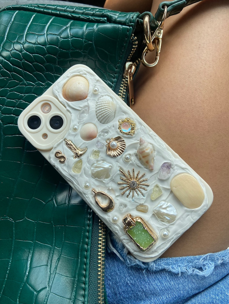 May include: White iPhone case with a seashell and pearl design. The case has a variety of seashells, pearls, and other beach-themed embellishments.