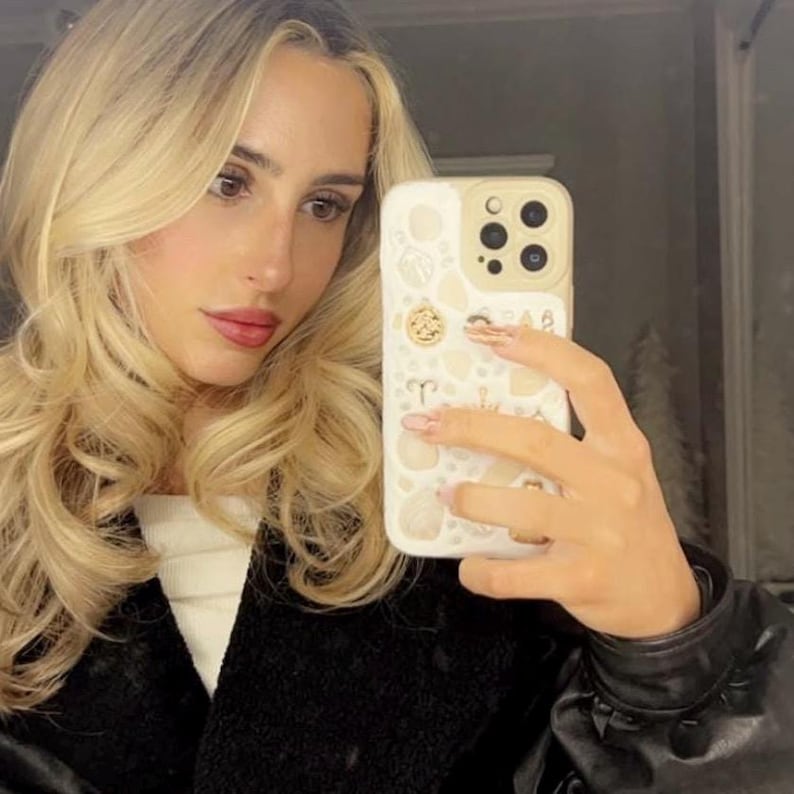 May include: A woman with long blonde hair is taking a selfie with a white iPhone case with gold accents and a variety of designs. The case has a gold zodiac symbol, a gold crown, and other gold accents.