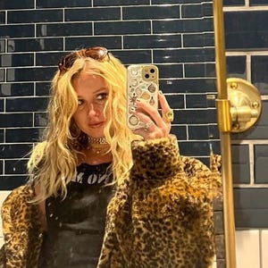 May include: A woman wearing a leopard print faux fur jacket, a black tank top with the text "M ON", and sunglasses. She is holding a white iPhone with a colorful sticker design.