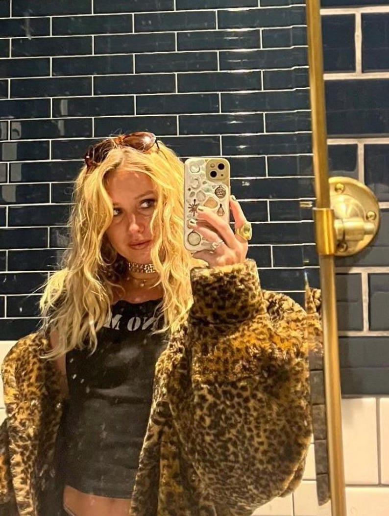 May include: A woman wearing a leopard print faux fur jacket, a black tank top with the text "M ON", and sunglasses. She is holding a white iPhone with a colorful sticker design.