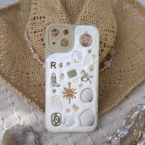 May include: White phone case with a variety of small charms and embellishments, including seashells, a star, a horseshoe, and a cowboy boot. The case has a clear camera lens cover and the text "Pro Camera" printed on it.
