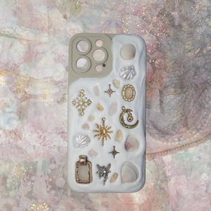 May include: A white phone case with a variety of gold and silver charms, including seashells, stars, and a cross. The case is decorated with a white, textured background.