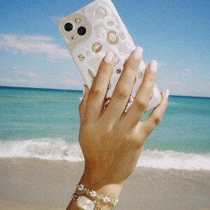 May include: A white iPhone case with a seashell and pearl design. The case is being held by a hand wearing a gold watch and a white and green beaded bracelet.