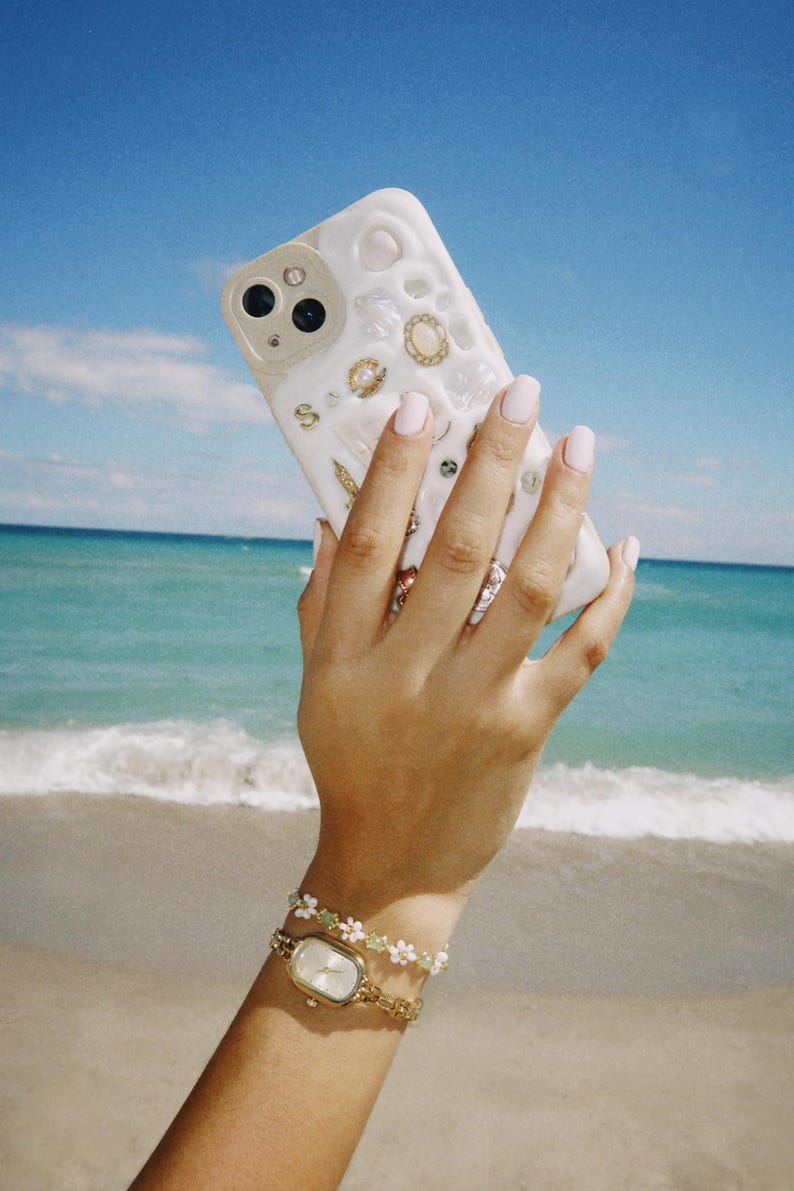 May include: A white iPhone case with a seashell and pearl design. The case is being held by a hand wearing a gold watch and a white and green beaded bracelet.
