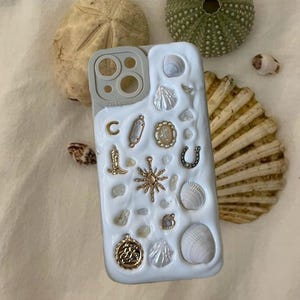 May include: White iPhone case with a decorative design featuring seashells, charms, and a gold sun. The case has a clear camera lens cover with three circles. The text "Pro Camera" is printed on the case.