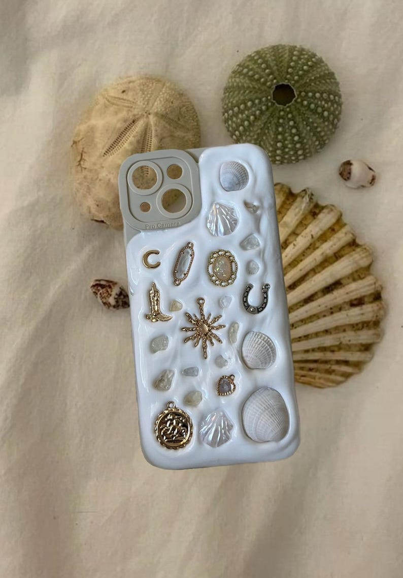 May include: White iPhone case with a decorative design featuring seashells, charms, and a gold sun. The case has a clear camera lens cover with three circles. The text "Pro Camera" is printed on the case.
