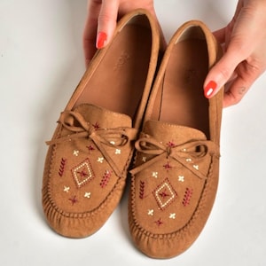 Może przedstawiać: Brązowe zamszowe mokasyny z haftowanymi wzorami w romby i kwiaty w kolorze czerwonym i białym. Buty mają kokardę na górze i są zaprojektowane z myślą o komforcie i stylu.