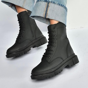 May include: Black combat boots with a lace-up design and a side zipper. The boots have a thick, lug sole and are made of a matte material. The boots are worn with blue jeans.