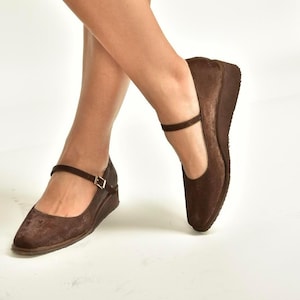 May include: Brown wedge heel shoes with a strap and buckle closure. The shoes have a textured appearance and a rounded toe. The wedge heel provides lift and support. The shoes are designed for comfort and style.