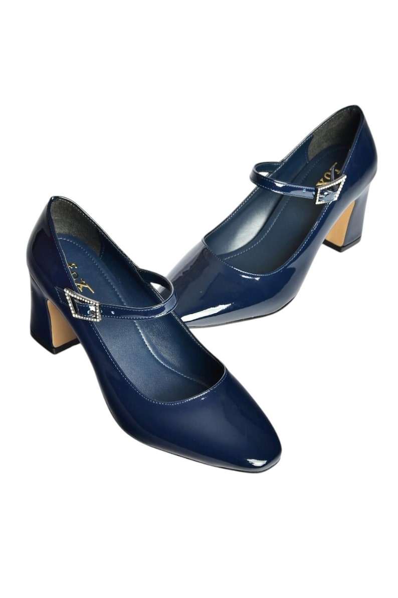 May include: A pair of navy blue patent leather Mary Jane heels. The shoes have a rounded toe, a strap across the top with a decorative buckle, and a block heel with a gold-colored base. The heels are approximately 2.5 inches high.