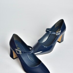 May include: A pair of navy blue patent leather Mary Jane heels. The shoes have a pointed toe, a strap across the top with a decorative buckle, and a block heel. The heels are approximately 3 inches high.