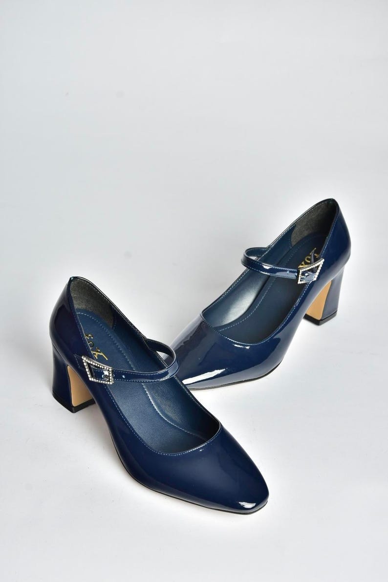 May include: A pair of navy blue patent leather Mary Jane heels. The shoes have a pointed toe, a strap across the top with a decorative buckle, and a block heel. The heels are approximately 3 inches high.