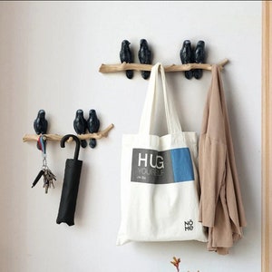May include: Wall-mounted coat hooks with black bird ornaments on wooden branches. A white tote bag with the text "HUG YOURSELF" hangs below the hooks, along with a beige jacket, keys, and a black umbrella. The hooks are fixed to a white wall.