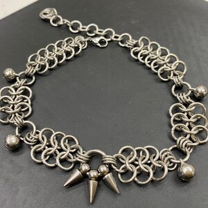 May include: A silver chain necklace with a chainmail design and silver ball accents. The necklace has a pointed spike detail at the centre.