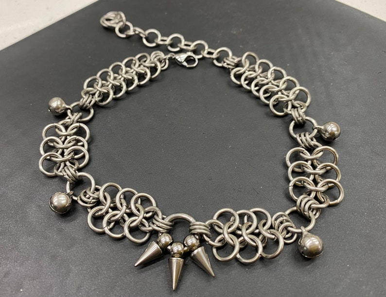 May include: A silver chain necklace with a chainmail design and silver ball accents. The necklace has a pointed spike detail at the centre.