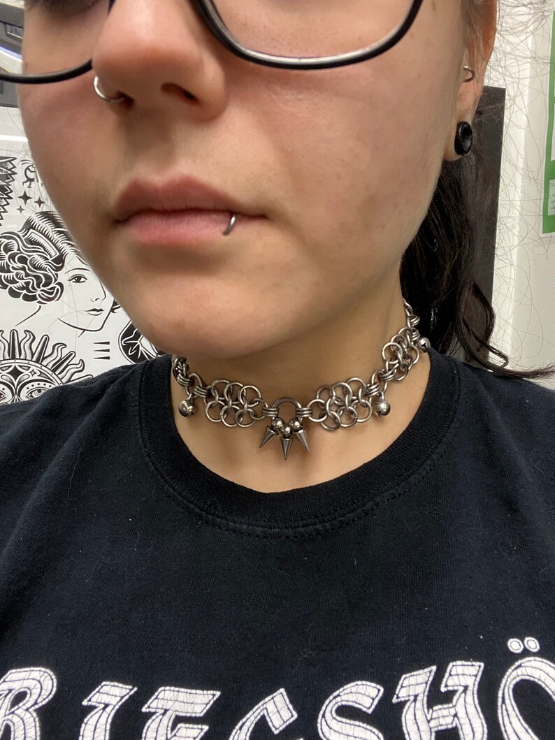 May include: A silver chain choker necklace with a spiked pendant and small silver balls. The necklace is worn by a person wearing a black shirt.