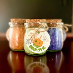 May include: Three small glass jars with cork stoppers, filled with orange, green, and purple contents. The center jar has a tag that says "Happy Halloween" and "Green Apple" with a pumpkin and spiderweb design. The jars are on a reflective surface.