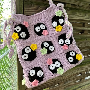 May include: A lavender crocheted tote bag with black and white cartoon faces and colorful flower accents.