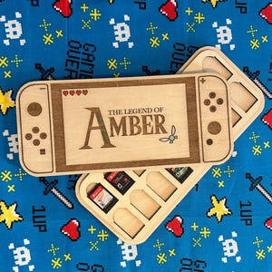 Könnte beinhalten: Holz-Aufbewahrungsbox in Form einer Spielkonsole mit dem Text "THE LEGEND OF AMBER" und einem Spielkartenhalter. Die Box ist aus hellem Holz mit einem dunkelbraunen Rand gefertigt. Der Hintergrund ist ein blauer Stoff mit pixeligen Spielmustern.