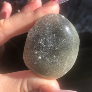 Peut inclure: Une pierre précieuse lisse, vert clair et gris, avec une texture mouchetée et scintillante. La pierre est de forme ovale et a une finition polie.