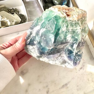 May include: A large, green and blue fluorite crystal with a rough, natural texture. The crystal has a variety of shades of green and blue, and some areas are translucent.
