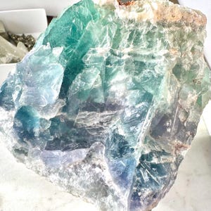 May include: A large, rough-hewn piece of fluorite crystal with a mix of green, blue, and white colors. The crystal has a natural, unpolished look.