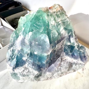 May include: A large, rough-hewn fluorite crystal with a mix of green, blue, and white hues. The crystal has a natural, unpolished appearance.