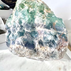 May include: A large, green, blue, and white fluorite crystal cluster. The crystal has a rough, natural texture and is displayed on a clear acrylic stand.