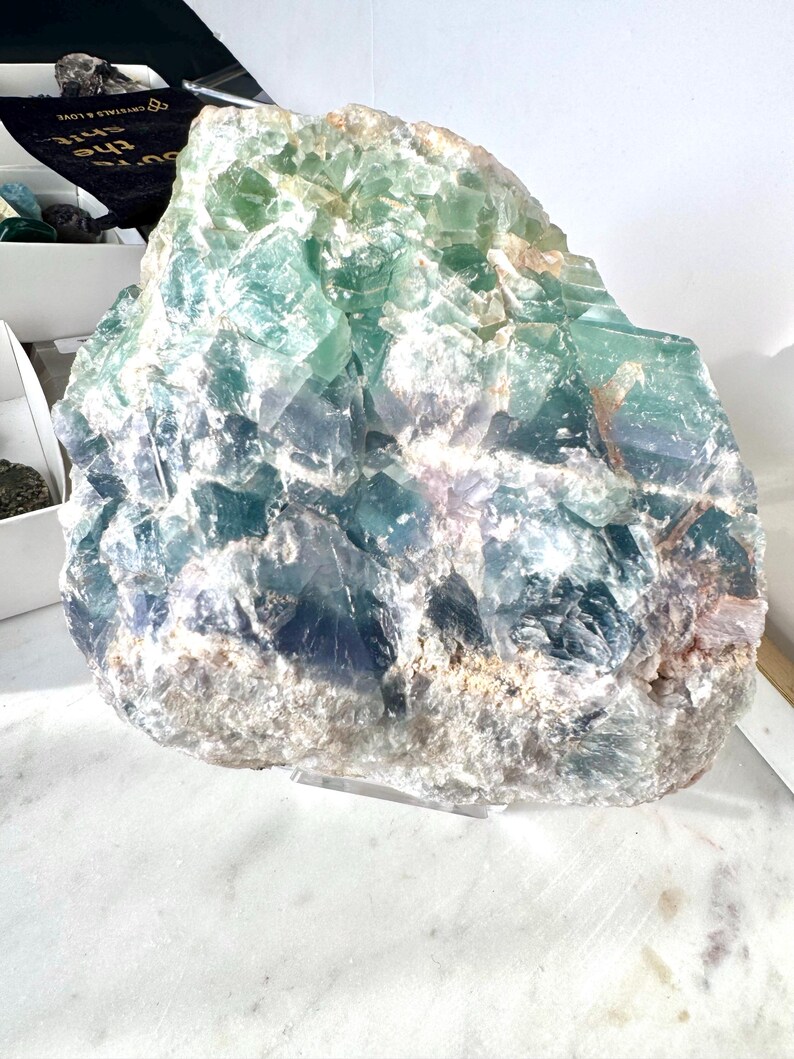May include: A large, green, blue, and white fluorite crystal cluster. The crystal has a rough, natural texture and is displayed on a clear acrylic stand.