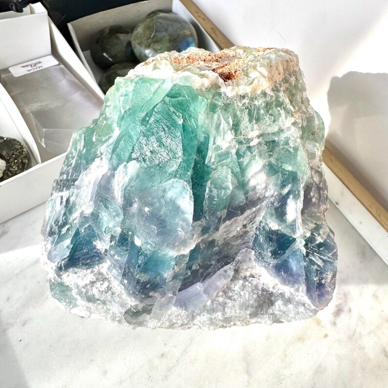May include: A large, rough-hewn piece of green and blue fluorite crystal. The crystal has a natural, unpolished surface and shows a variety of shades of green and blue.