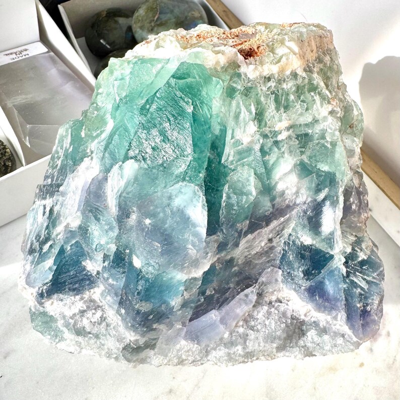 May include: A large, rough-hewn green and blue fluorite crystal with a white, cloudy interior. The crystal has a natural, irregular shape and is a beautiful example of this mineral.